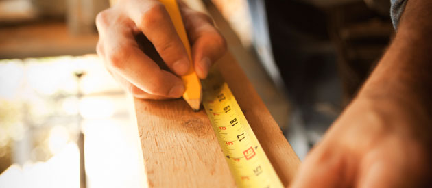 Ein Schreiner ist auf einem langen Stück Holz mit einem Massstab eine Strecke am abmessen und zeichnet sie mit einem gelben Bleistift ein.